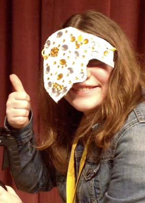 A young woman gives a “thumbs up” while wearing a mask made of gold and silver jewels on a white background that covers half of her face.