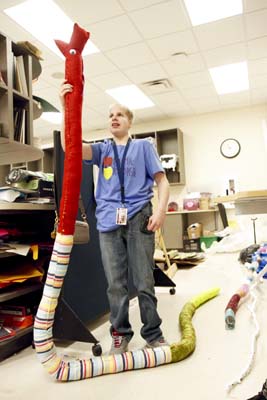 Young man holds up long stuffed snake twice as long as he is, red fabric and googly eyes for head, striped fabric for body, and green fabric for tail.