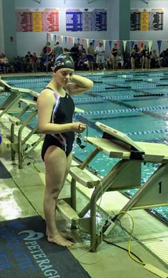 Photo of a teenage girl in a black swimsuit standing next to the diving blocks of an indoor swimming pool. She has swim goggles in her hands.