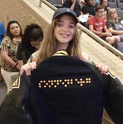 A smiling teenage girl with long hair holds a navy letter jacket with yellow dots on the back which spell “Sonnenberg” in braille. 