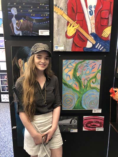 Scout stands in front of a display of artwork. There are four large paintings and two small ones. Scout's is a multicolored painting of a tree.