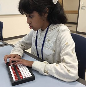 A young girl with dark hair sits at a table. Her hands are moving beads on an abacus.
