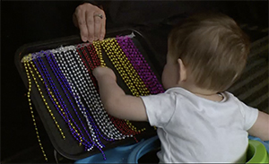 An infant boy sits facing a multicolored beaded curtain on a black slant board holding onto a gold mardi-gras strand of beads.