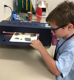 A male student with dark blue glasses feeds a piece of thermaform paper with drawings of different shapes into a PIAF tactile graphic maker.