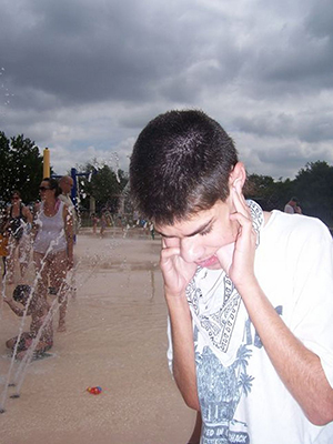 A young man stands near others who are enjoying a splash pad. He holds his ears closed with his fingers while his thumbs are inserted all the way into his mouth between his teeth, palms up.