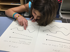 A female student uses her right hand to examine a tactile graphic containing raised line forms and braille.