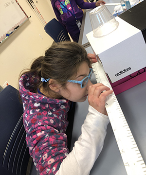 A female student with blue glasses uses a tactile ruler to measure a box.