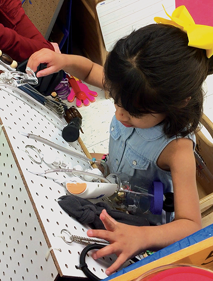 Una niña pequeña está sentada delante de una pizarra de clavijas y usa ambas manos para explorar objetos colocados allí, incluso cadenas, artículos con orificios y frascos con tapas.