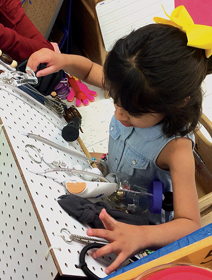 A young girl sits in front of a piece of peg-board and uses both hands to explore objects attached to it, including chains, items with holes, and containers with tops.