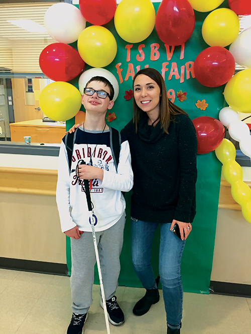 A student stands with an adult in front of a green backdrop topped with red and yellow balloons.