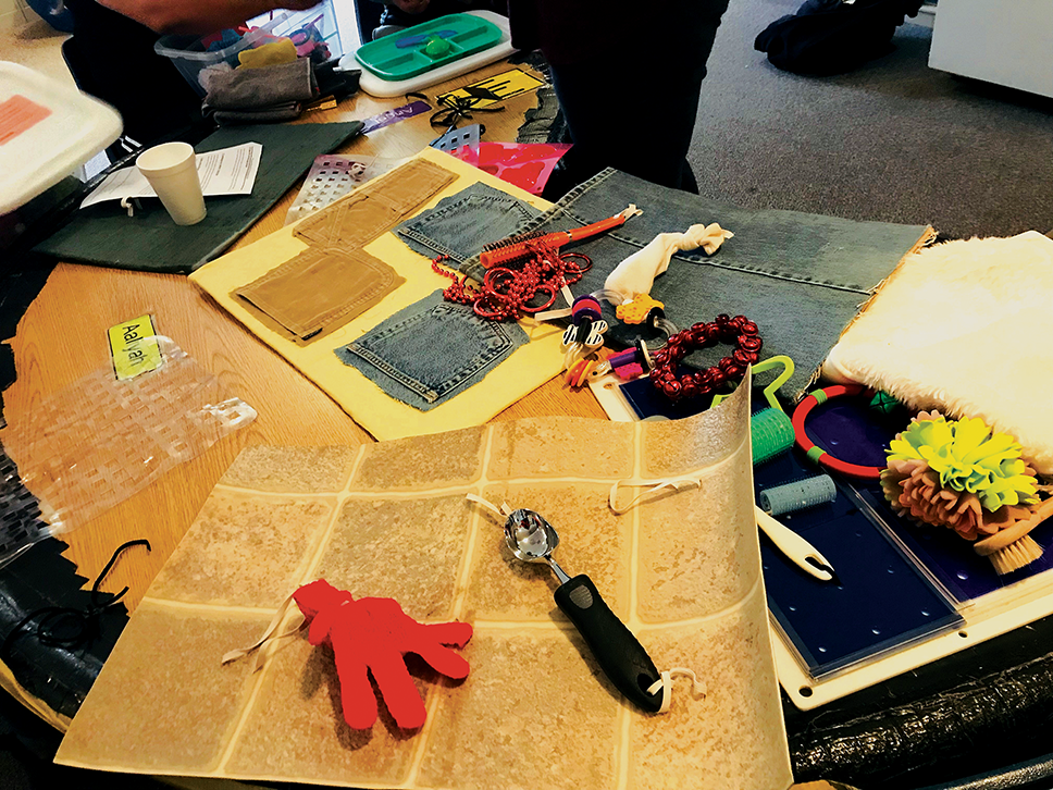 A variety of tools and materials are displayed on a table, including an ice cream scoop, a hair roller, denim fabric, beads, and a red glove.