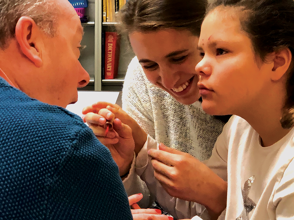 Una niña pequeña y su maestra pintan de rojo las uñas de un señor. La niña y el hombre mantienen contacto visual mientras la maestra sonríe.