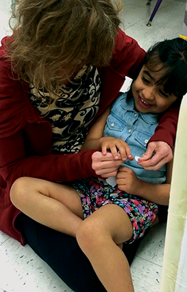 A young girl sits in an adult’s lap, laughing. She holds a piece of paper in one hand and explores the actions of the adult’s hands with the other.