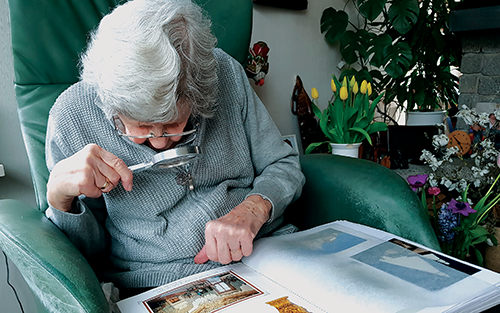 A gray-haired woman holds a magnifier in her right hand to help her read the book in her lap.