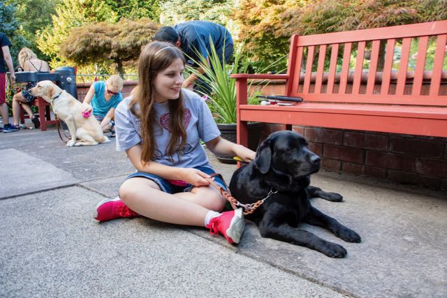 A young student with long brown hair sits outside hugging a black lab.