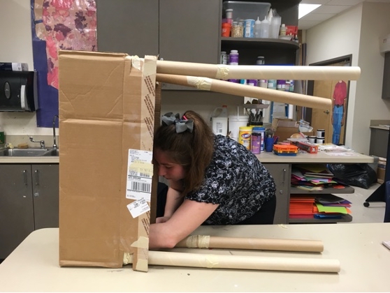 A student works with a cardboard box and cardboard tubes on a classroom table.