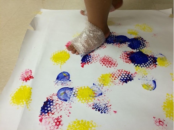 A child with one foot wrapped in bubble wrap stands on a large piece of white paper.