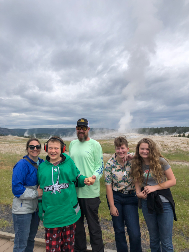 A family of 5—two parents and three children—stands in front of an erupting geyser.