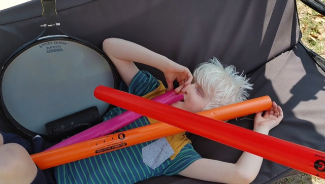 A young student lies on a bench swing. The student’s body is in contact with a paddle drum, a small speaker, and colorful tuned percussion tubes.