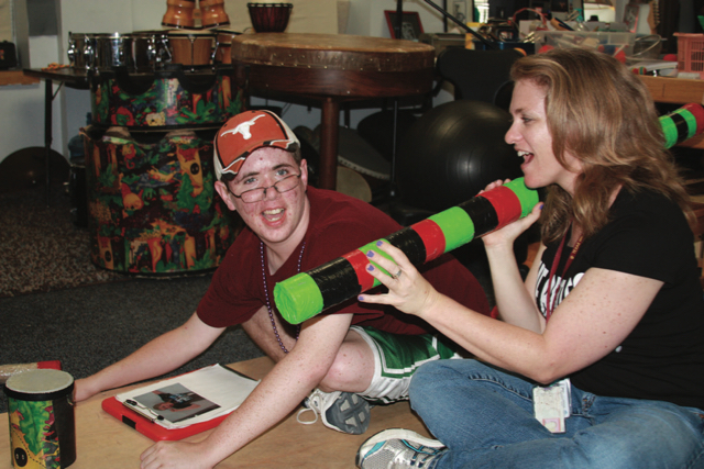 A teenage student sits cross-legged on a resonance board with TSBVI music instructor who is holding a large colorful rainstick.