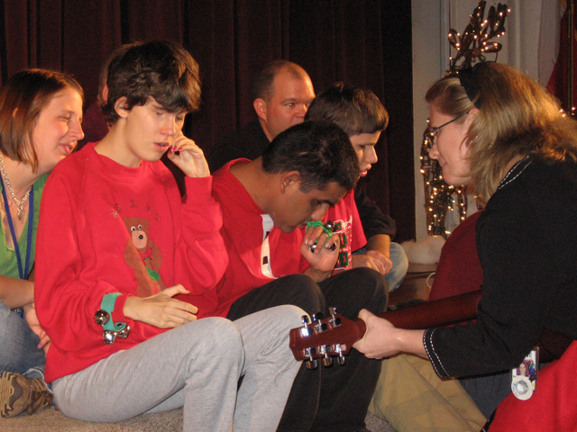 Three teenage students hold jingle bells and sit on a stage with two staff members while their music teacher plays a guitar. 