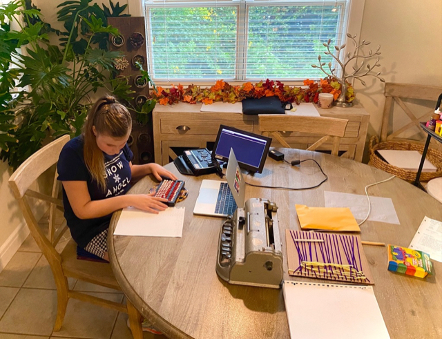 A student sits at a table with an abacus, a braille notetaker with external monitor, a laptop, a Perkins brailler, and art materials.