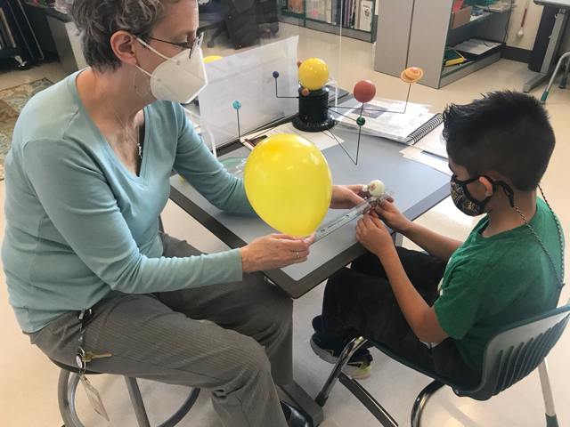 An instructor conducts a science experiment using a ping pong ball, golf tee, marshmallow, and balloon to represent the earth, moon, and sun.