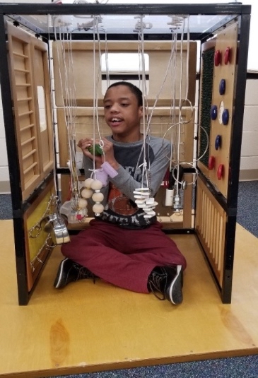 A student sits up in an adult-sized Little Room. Objects hang down on long elastic so the student can play with them at chest level.