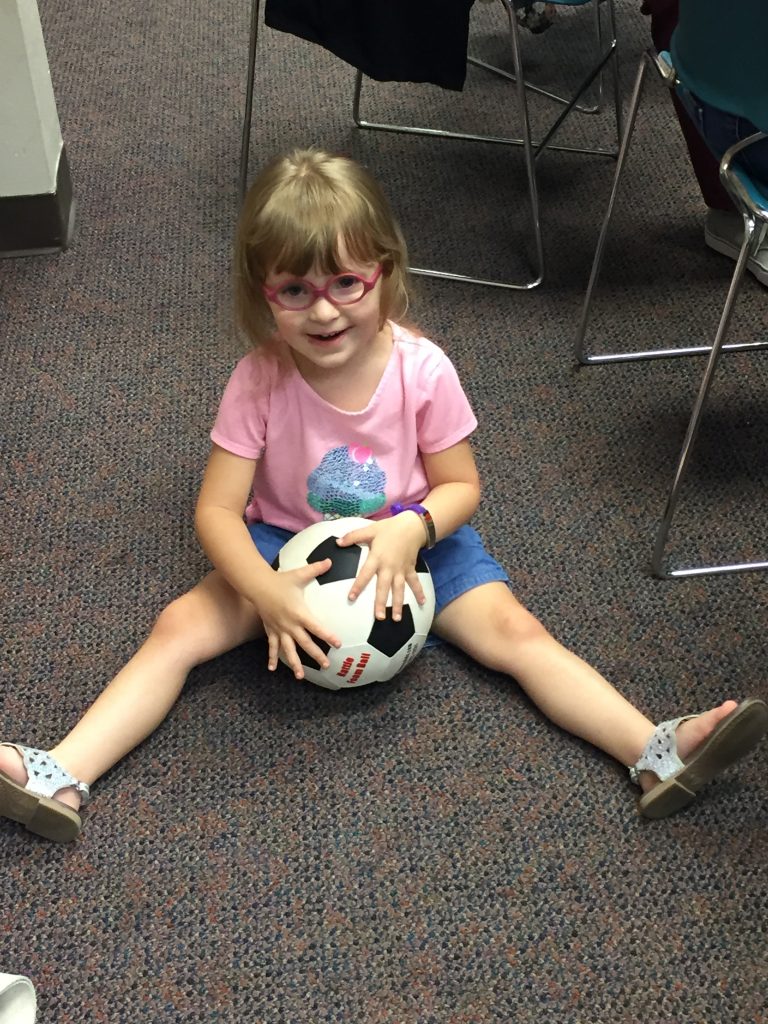 A toddler with pink glasses, smiles while sitting on the ground and holding a ball.