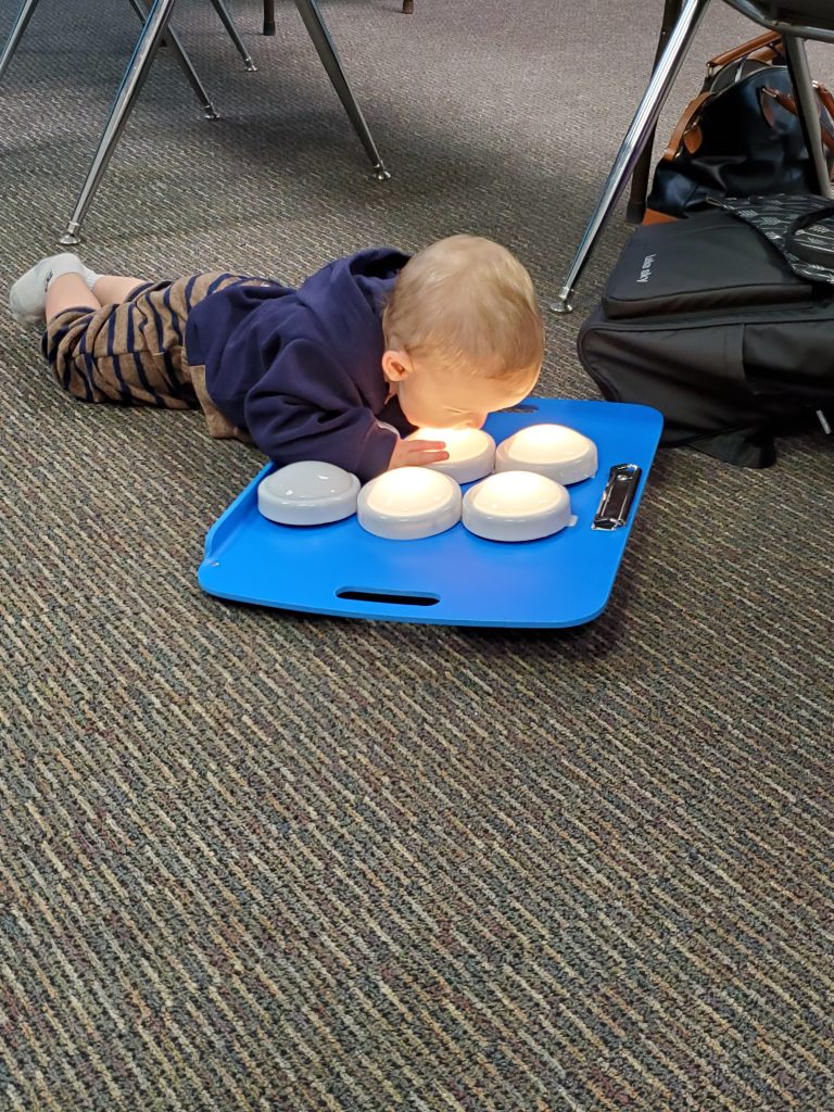 A young toddler lies on the ground and looks at a display of 6 bright lights in the configuration of a braille cell.