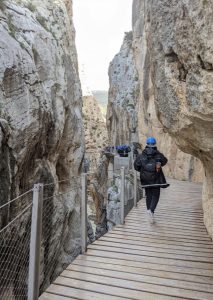 Una joven adulta con abrigo, mochila y casco camina por un estrecho sendero de madera entre peñascos rocosos.