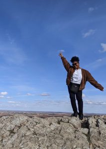 Una joven adulta con gafas de sol en lo alto de una montaña con los brazos en alto en señal de celebración.