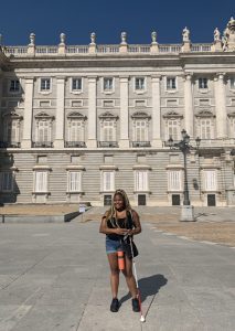 Una joven adulta con un bastón en la mano, parado frente a un gran edificio majestuoso.