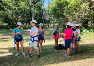 Seven students wearing white helmets stand together in front of tall wooden obstacles and a net.