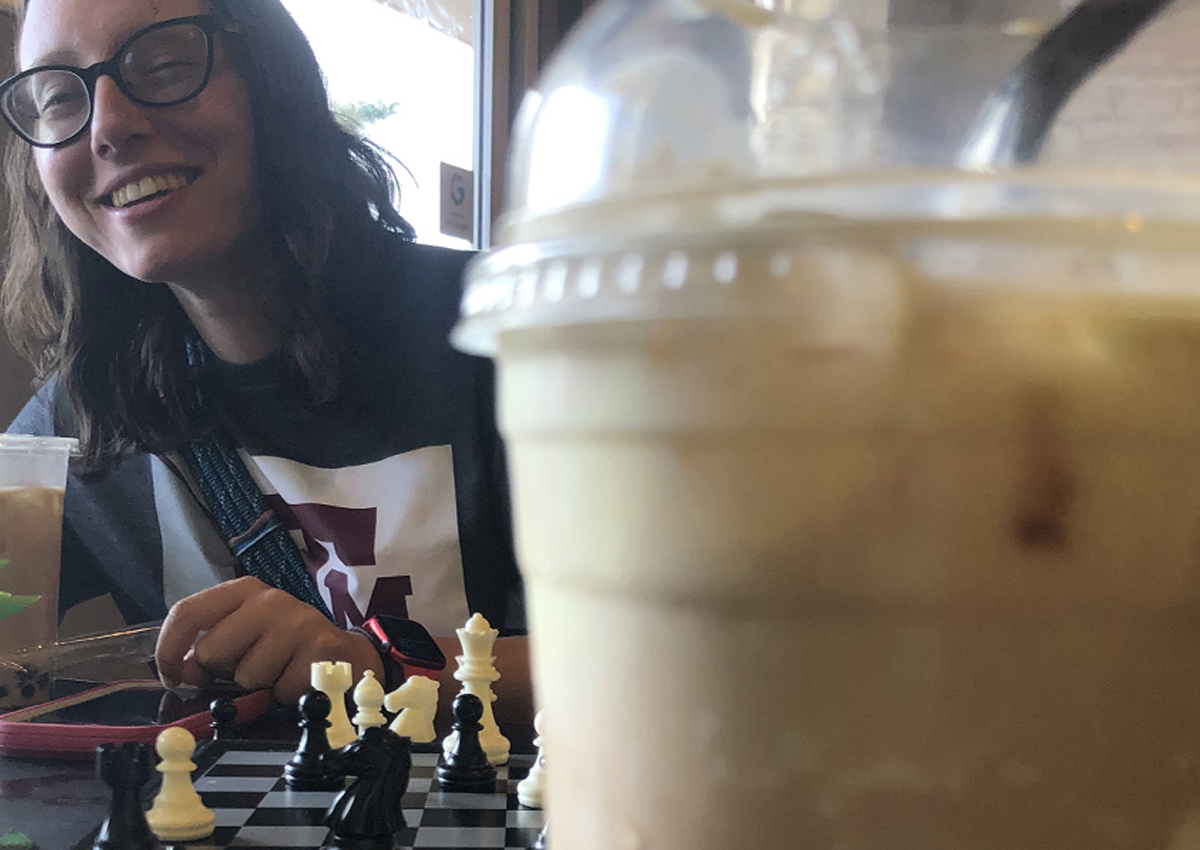A young adult with glasses sits at a table with an iced drink and a game of chess.