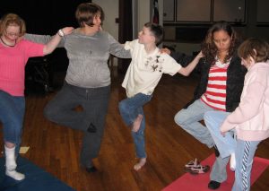 An adult, two staff, and three students stand on yoga mats with their arms on each other’s shoulders, each with one leg bent up to touch the k