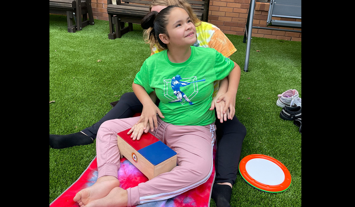 An adult sits behind a smiling adolescent and places her hands under the student’s to make contact with a rectangular drum in front of them. They are sitting on astro-turf outdoors.