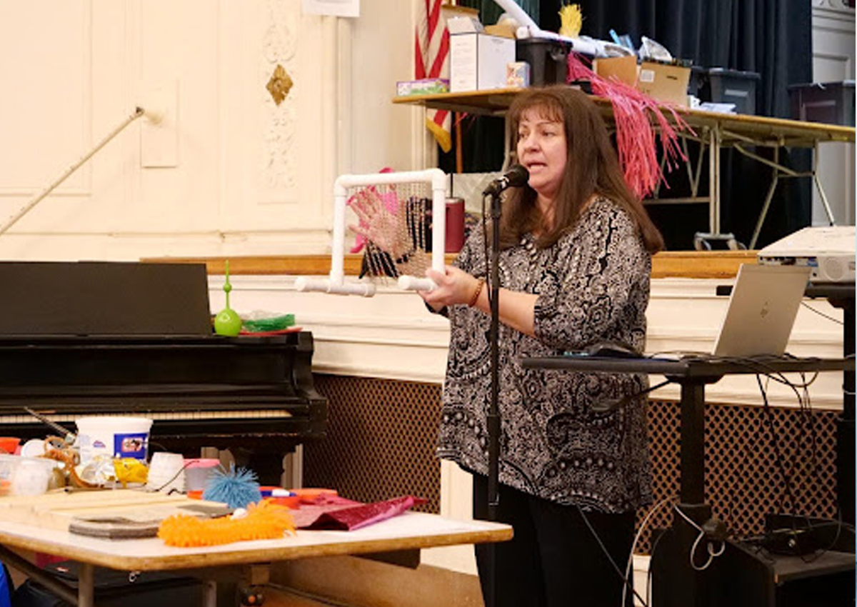 An adult stands behind a table with colorful multi-textured objects and speaks into a microphone on a stand.
