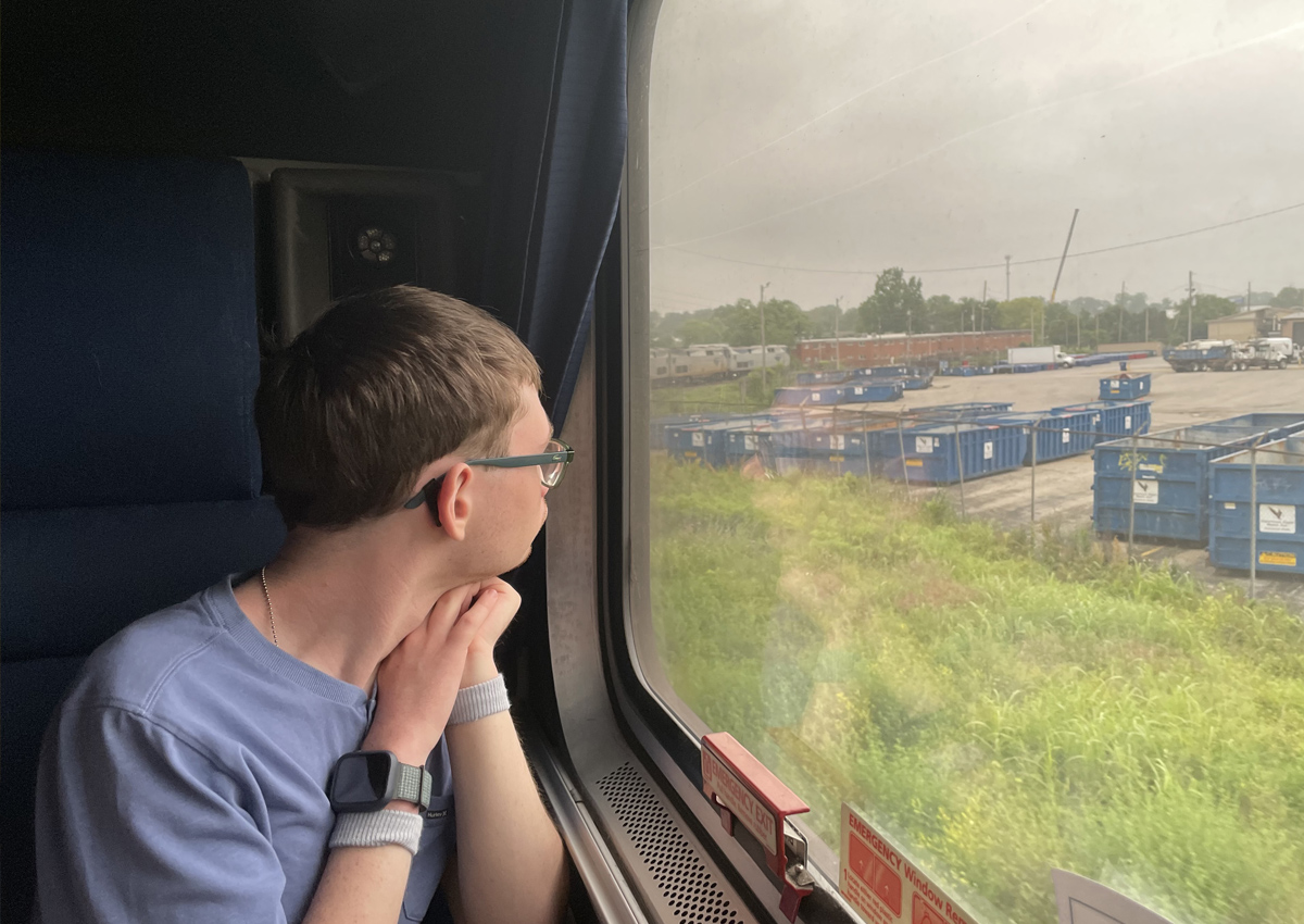 An adolescent looks out the window of a train with his hands clasped under his chin.