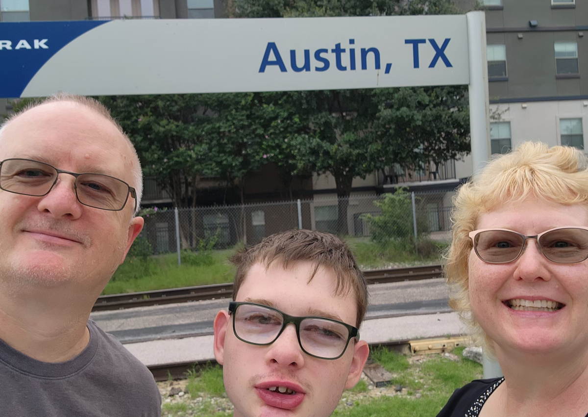 An adolescent and his mom and dad smile for a selfie in front of the Amtrak Austin, TX sign.