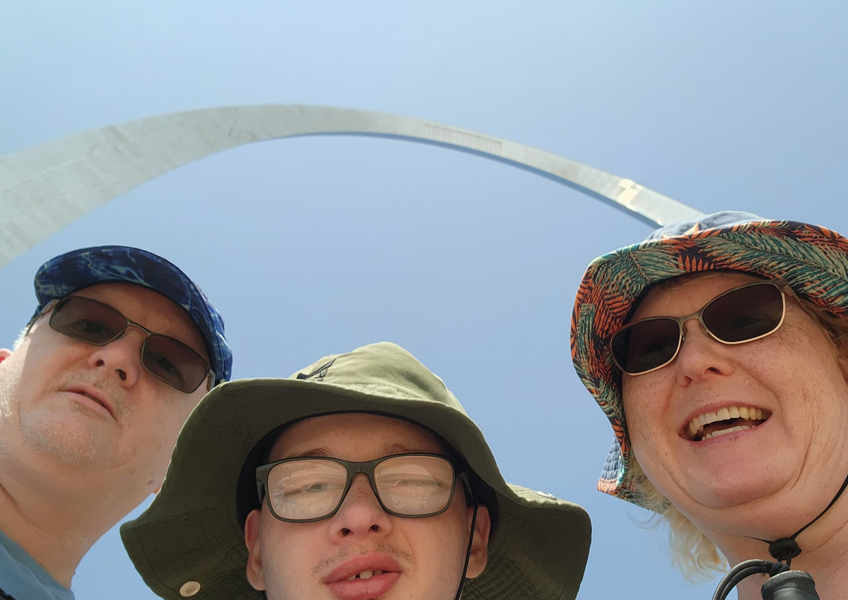 An adolescent and his mom and dad pose for a selfie while standing under the St. Louis Arch.