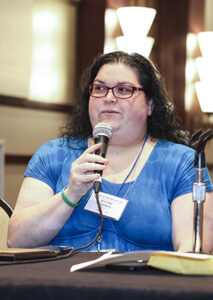 A woman with glasses and long brown hair sits at a table holding a microphone.