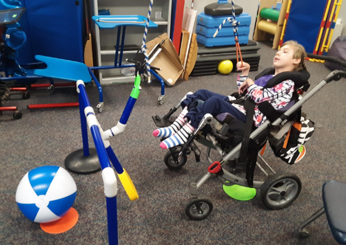 A student in a wheelchair uses a cord to pull a lever made of PVC pipe to hit a beach ball.