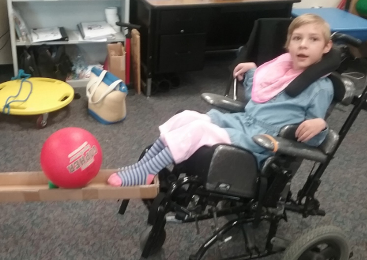 A student in a wheelchair uses his foot to kick a ball toward a bowling pin that is contained in a long rectangular box.