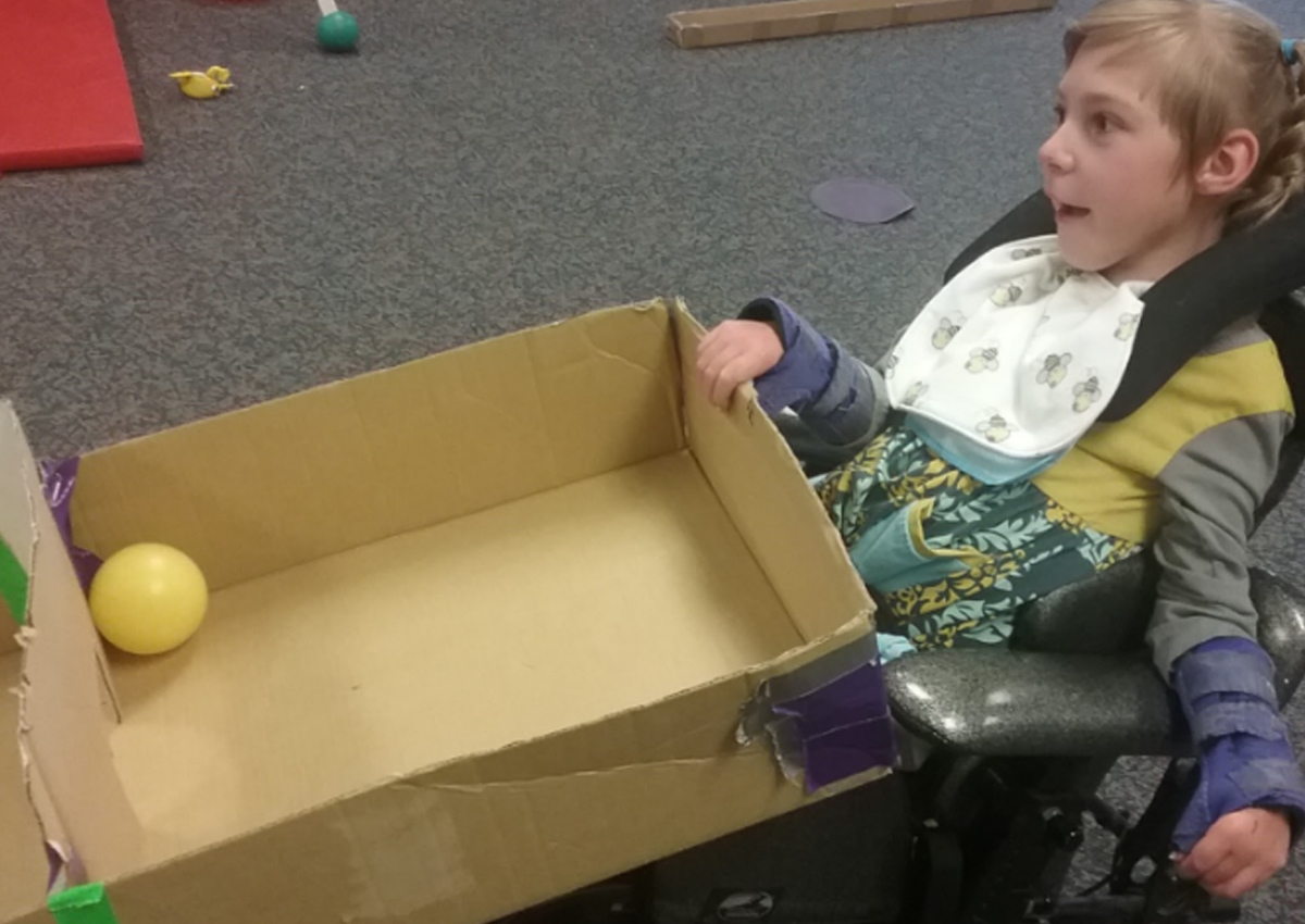 A student uses his right hand to tilt a cardboard box in order to move a ball toward a goal on the other side of the box.