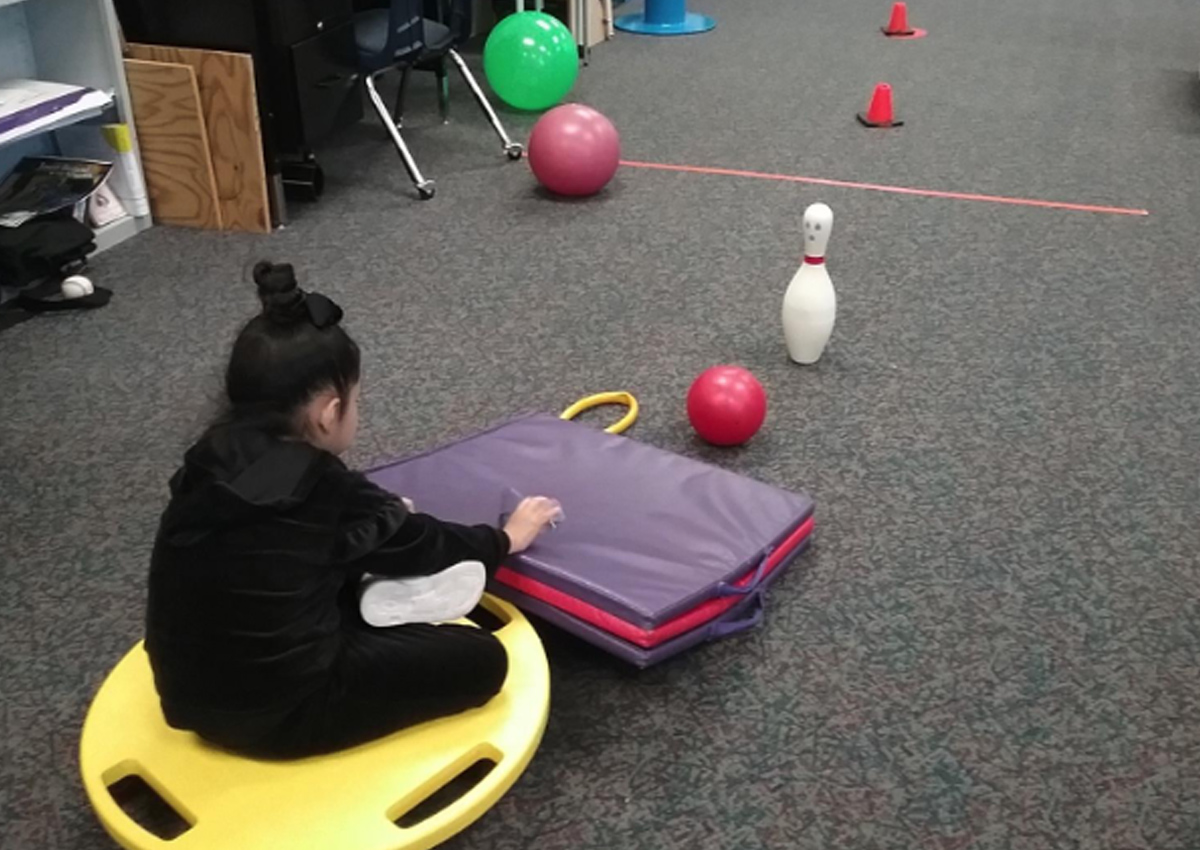 A student sitting on a sit-n-spin uses a folded and angled floor mat to hit a ball toward a bowling pin.