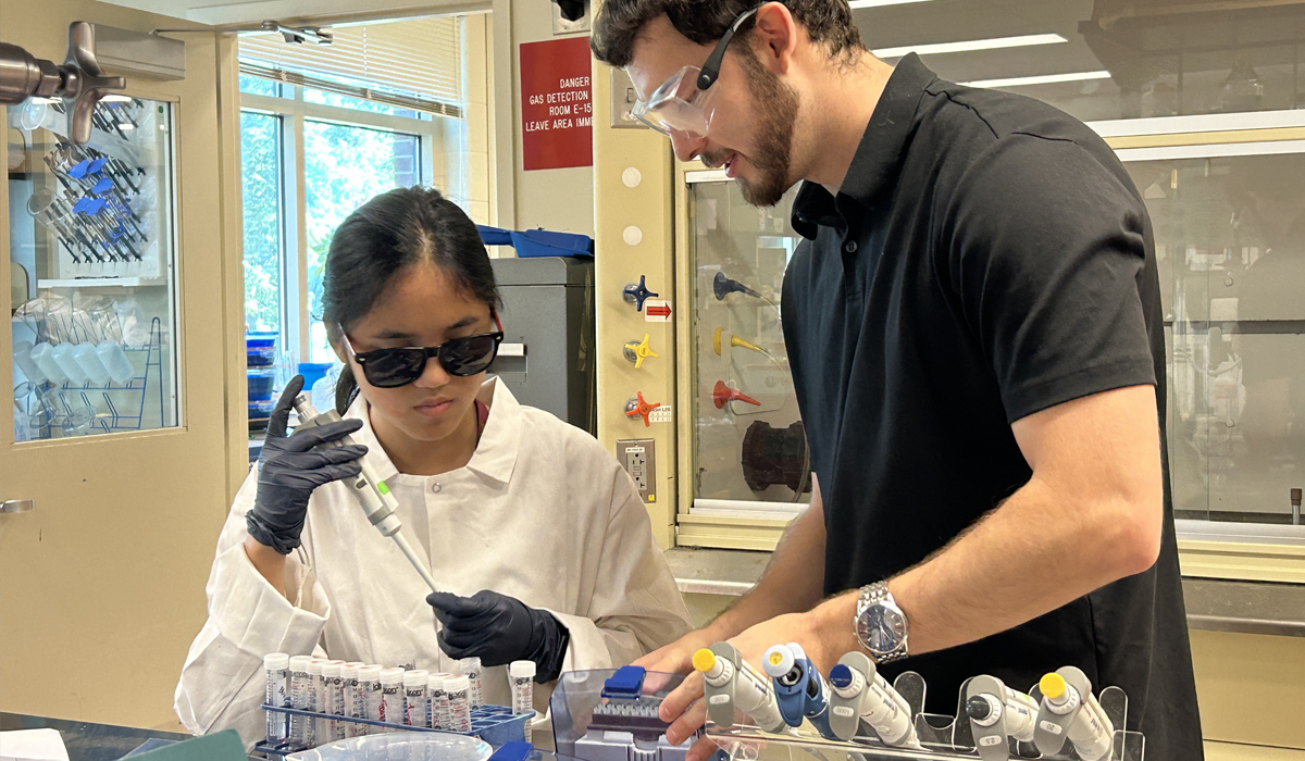 An adolescent in a lab coat holds a pipette to a test tube while an adult in safety goggles stands beside her in a chemistry lab.