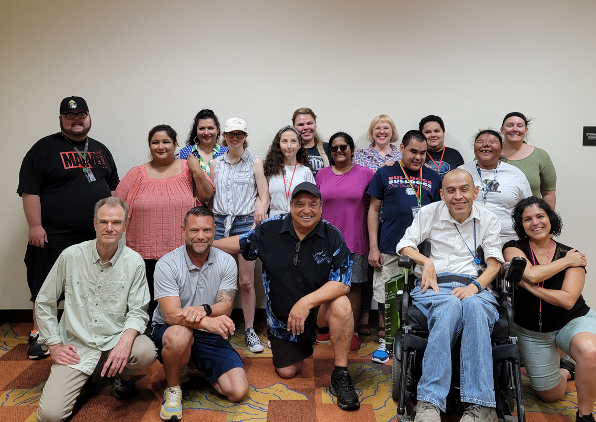 Two rows of people smile for a group photo. The front row is kneeling. One member is in a wheelchair.