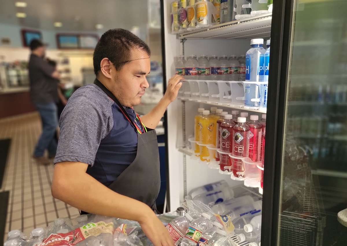 An adolescent wearing a black apron arranges items on a shelf in the refrigerated section of a convenience store.