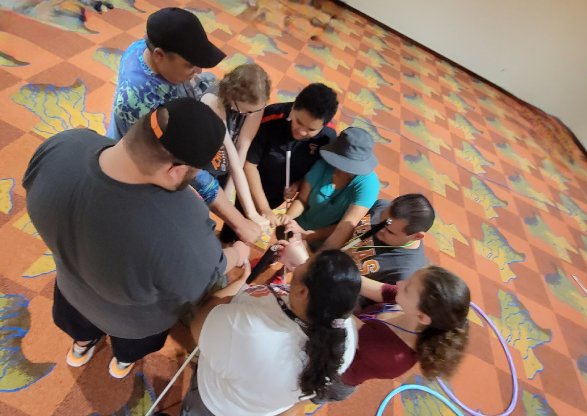 Eight students stand in a circle with their hands joined in the center.